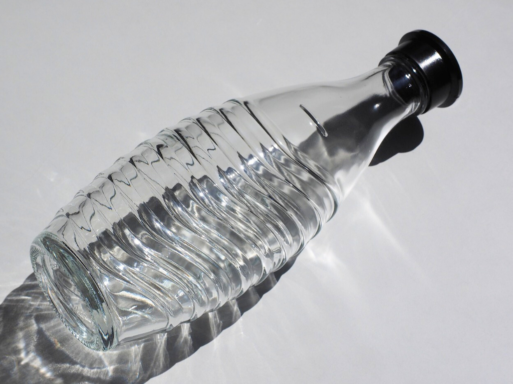 Eine leere, liegende Glasflasche mit spiralförmiger Rillenstruktur und einem schwarzen Verschluss liegt auf einem weißen Untergrund und wirft einen starken Schatten.