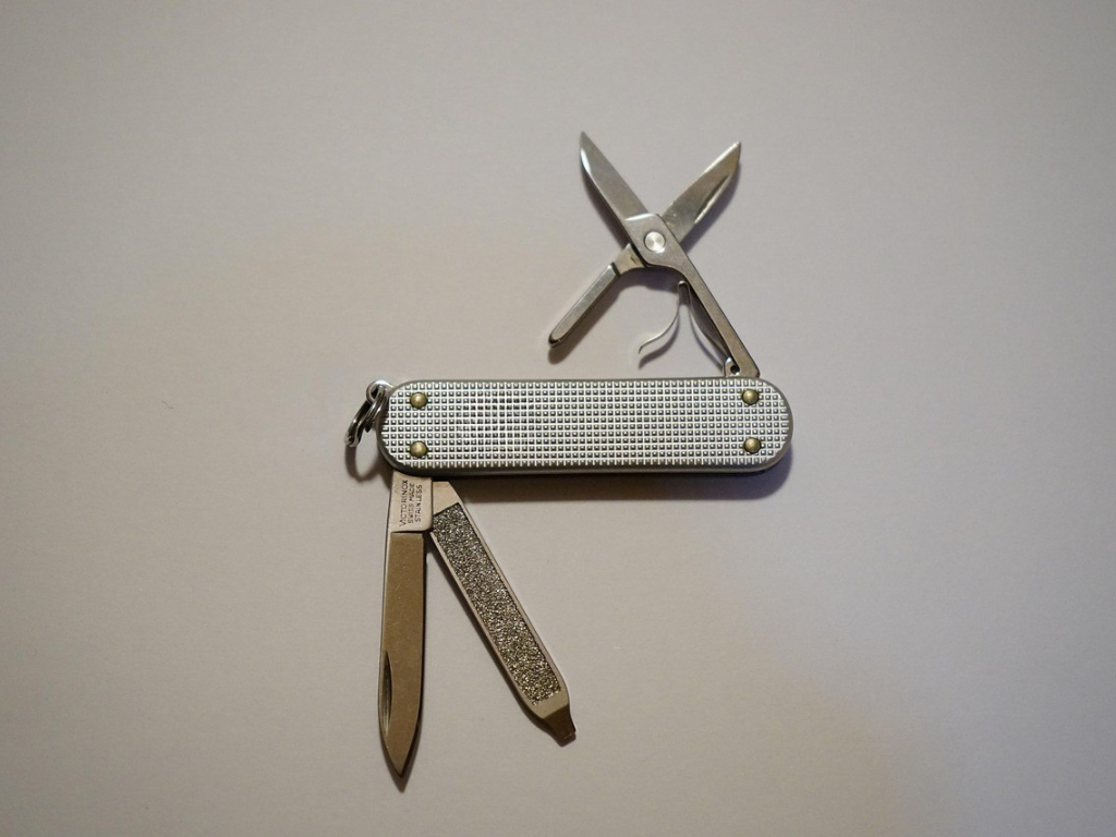 Ein silbernes Schweizer Taschenmesser mit aufklappbaren Werkzeugen, darunter eine kleine Klinge, eine Nagelfeile und eine kleine Schere, liegt auf einem hellgrauen Untergrund.