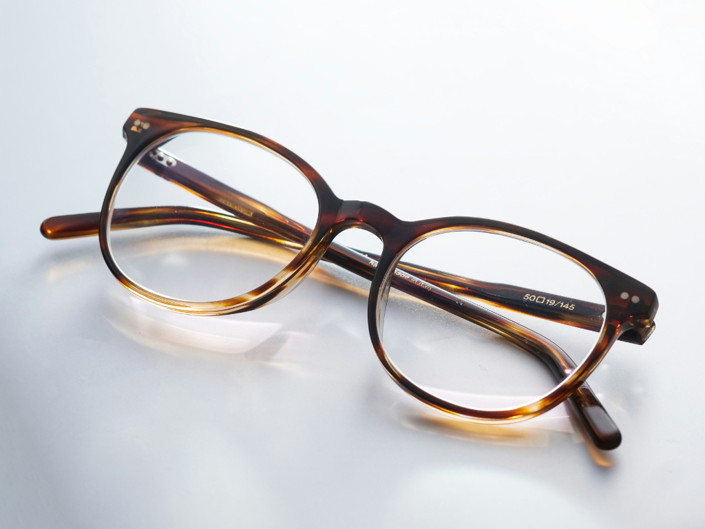 Eine Brille mit einem braun-gemusterten Rahmen und runden Gläsern liegt auf einer hellen, reflektierenden Oberfläche.