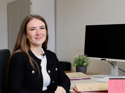Porträt einer Justizfachwirtin am Schreibtisch. Sie lächelt in die Kamera, vor ihr liegen Arbeitsunterlagen und im Hintergrund sind zwei Computerbildschirme zu sehen.