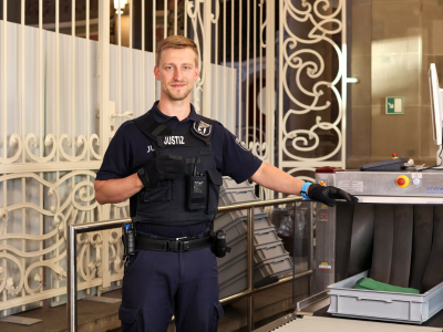 Ein Justizhauptwachtmeister in dunkelblauer Dienstkleidung steht neben einem Gepäckscanner im Eingangsbereich. Er trägt eine Schutzweste mit der Aufschrift 'JUSTIZ'.