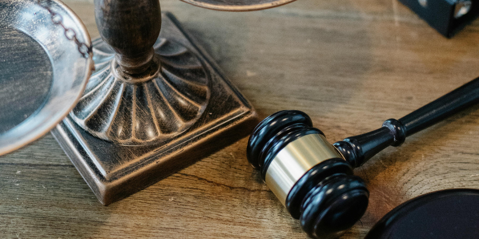 Nahaufnahme eines Richterhammers aus dunklem Holz, der neben dem verzierten Standfuß einer Waage auf einem Holztisch liegt.