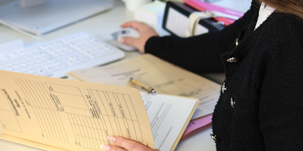 Nahaufnahme einer Person am Schreibtisch, die eine beige Akte in der Hand hält und gleichzeitig eine Computermaus bedient. Im Hintergrund sind weitere Arbeitsunterlagen sichtbar.