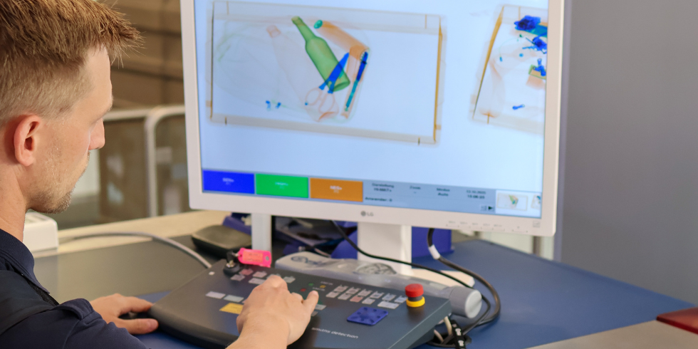 Ein Mitarbeiter an der Sicherheitskontrolle betrachtet einen Monitor, der Röntgenbilder von Gepäckstücken mit Flaschen und Scheren zeigt, und bedient dabei ein Steuerpult.