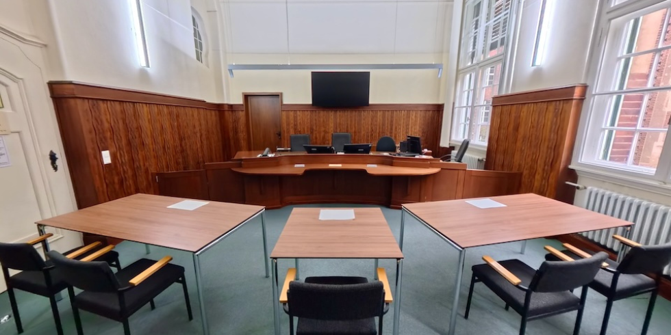 Weitwinkelaufnahme eines hellen Gerichtssaals. Im Vordergrund stehen Tische für die Prozessbeteiligten, im Hintergrund thront die Richterbank auf einem Podest vor einer holzvertäfelten Wand.