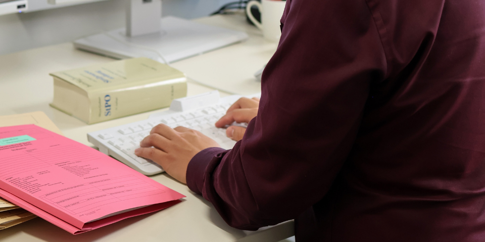 Rückansicht einer Person, die am Computer arbeitet. Im Vordergrund liegen eine rosa Akte und ein Gesetzbuch mit der Aufschrift 'StPO' auf dem Tisch.
