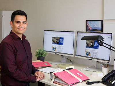 Porträt eines Rechtspflegers an seinem Arbeitsplatz. Er sitzt an einem Schreibtisch, auf dem dicke Aktenbündel, ein Gesetzbuch und zwei Monitore stehen.