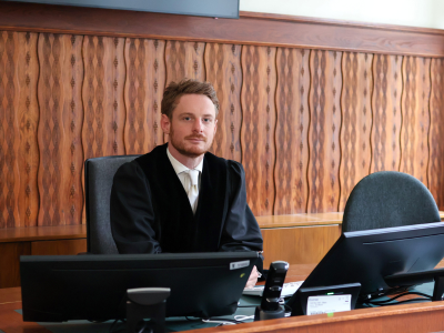 Ein Staatsanwalt sitzt an einem Tisch in einem Gerichtssaal. Er trägt eine schwarze Robe und eine weiße Krawatte; im Hintergrund ist eine wellenförmige Holzvertäfelung zu sehen.