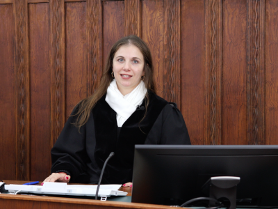 Eine Richterin sitzt in einem holzgetäfelten Gerichtssaal am Richtertisch. Sie trägt eine schwarze Robe mit weißem Halstuch und blickt in die Kamera.