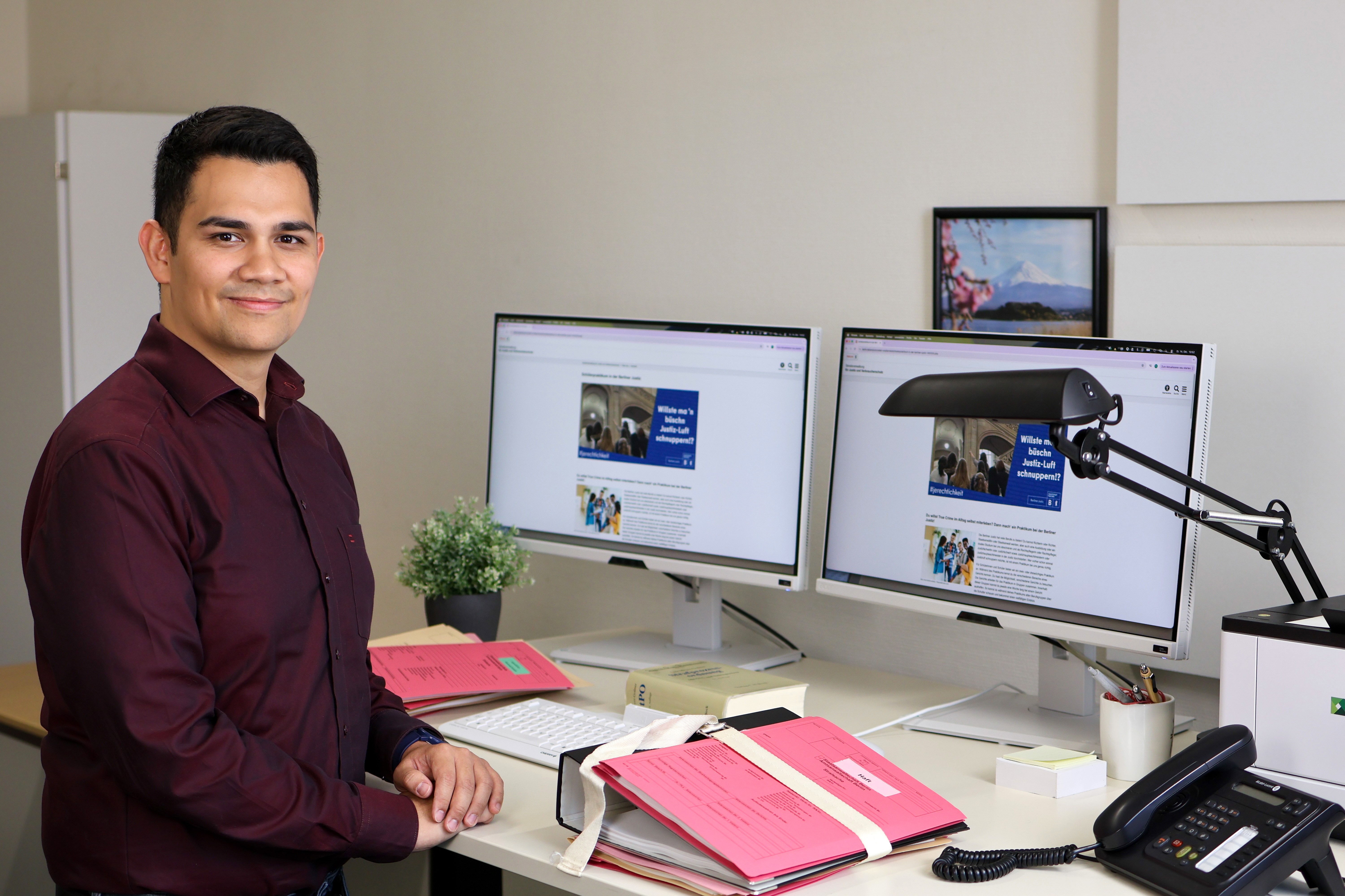 Porträt eines Rechtspflegers an seinem Arbeitsplatz. Er sitzt an einem Schreibtisch, auf dem dicke Aktenbündel, ein Gesetzbuch und zwei Monitore stehen.