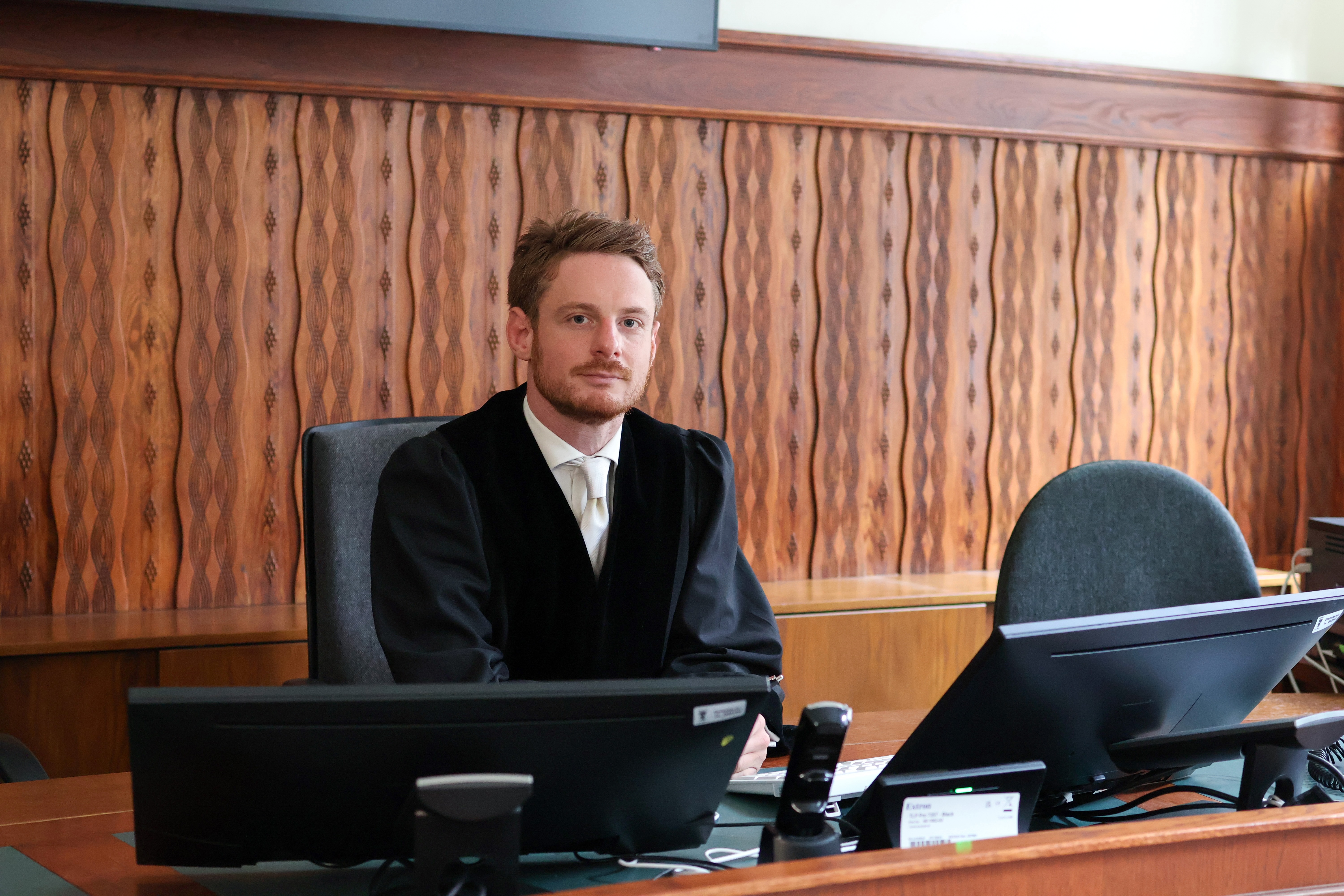 Ein Staatsanwalt sitzt an einem Tisch in einem Gerichtssaal. Er trägt eine schwarze Robe und eine weiße Krawatte; im Hintergrund ist eine wellenförmige Holzvertäfelung zu sehen.